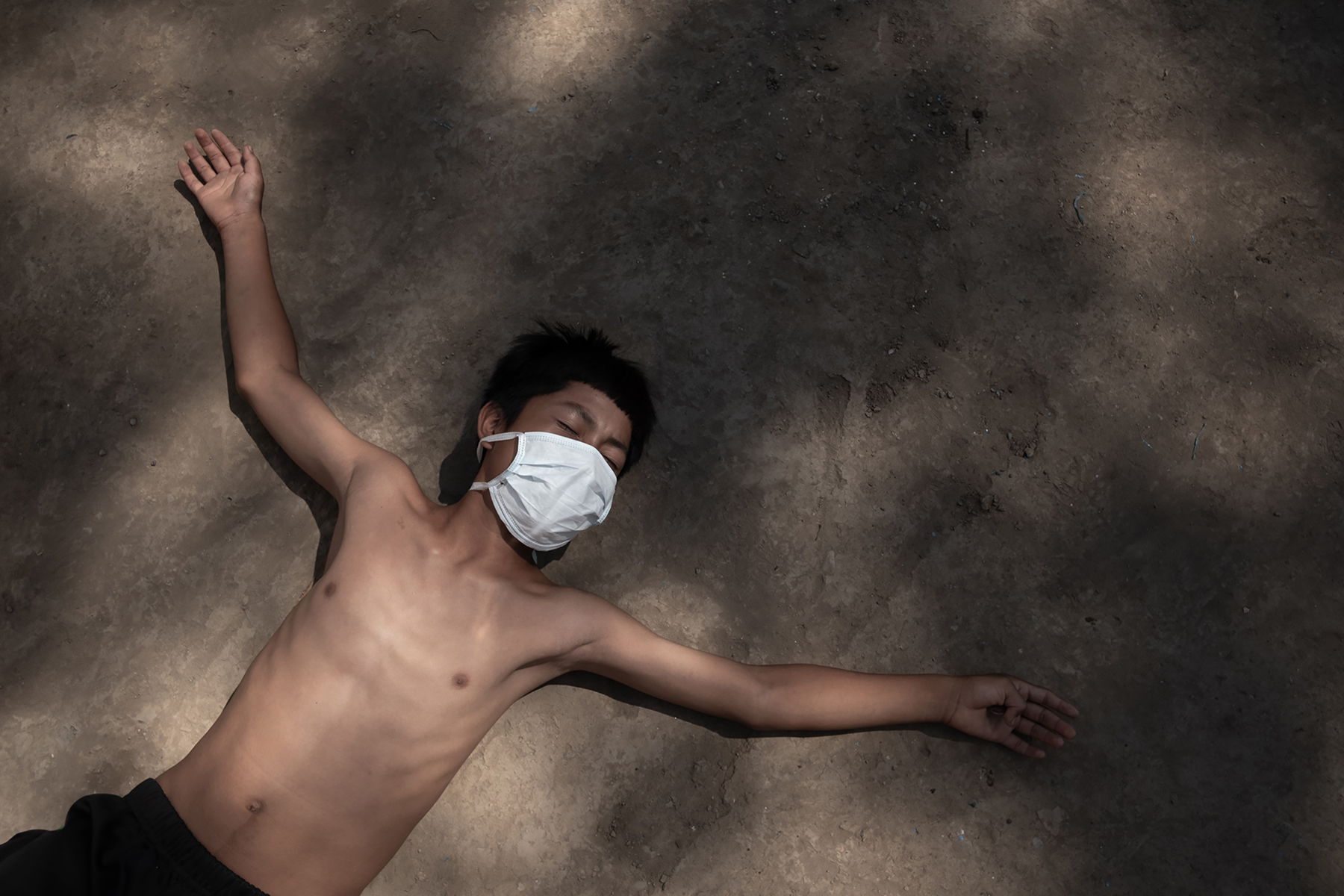 Image by Florence Goupil. Peru, 2020. A young boy lies down on the dirt ground with arms outstretched. He is wearing a white surgical mask, black shorts, and no shirt.