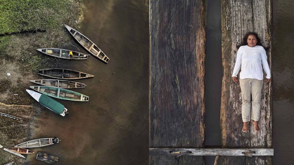 lilia rainforest defenders.jpg To the left, a series of empty wooden canoes stand by the margins of a river. On the right, Lilia Isolina Java Tapayuri lies down on a plank of wood, looking directly at the camera, captured from an aerial shot. She is wearing a white long sleeved shirt, khaki-colored pants, and no shoes.