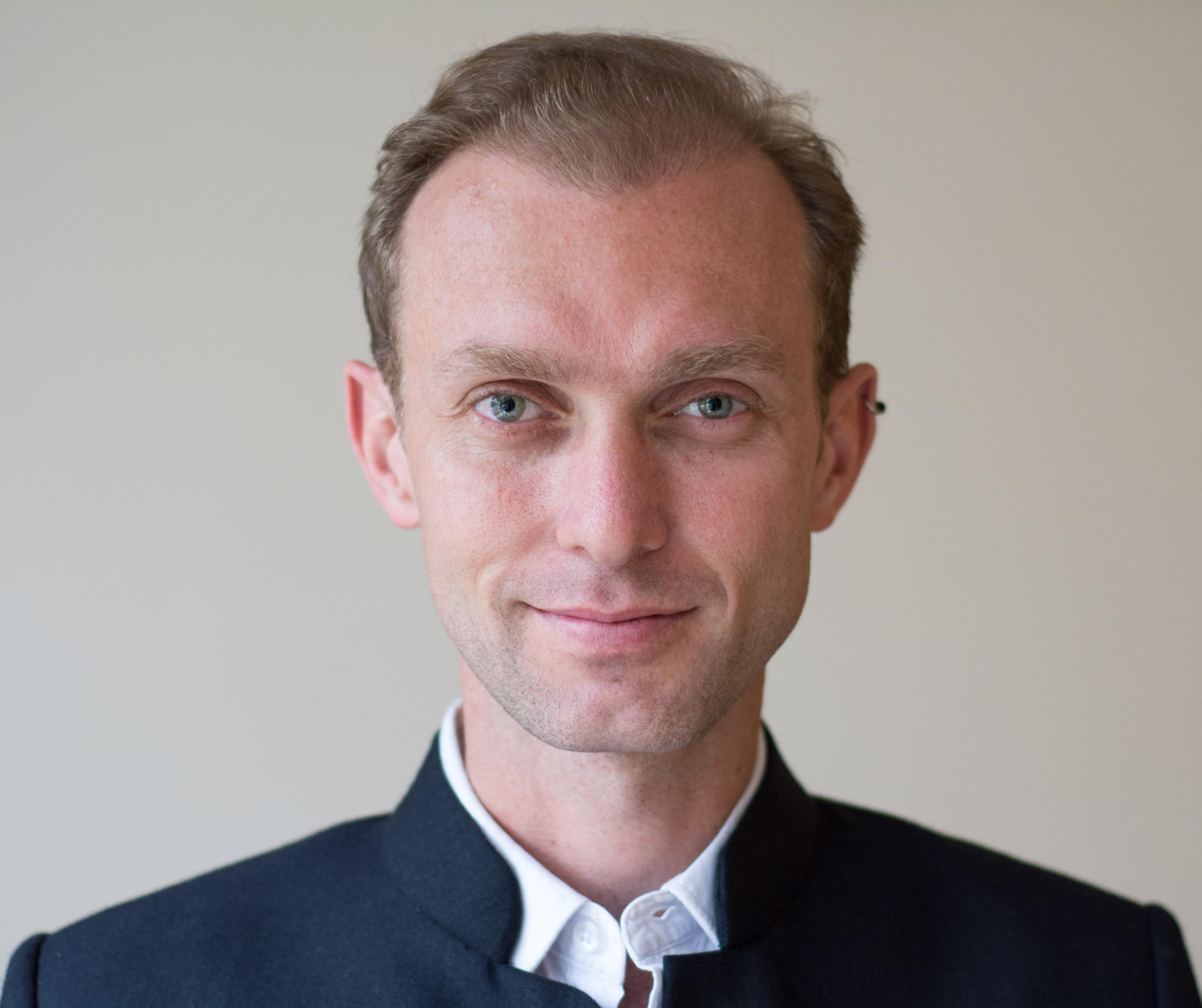 Sébastien Farcis A man wearing a white shirt and dark blue jacket. He has an ear piercing.