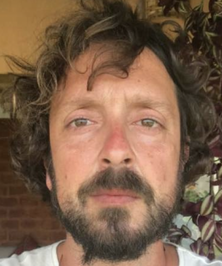 A man wears a white shirt and has brown curly hair.