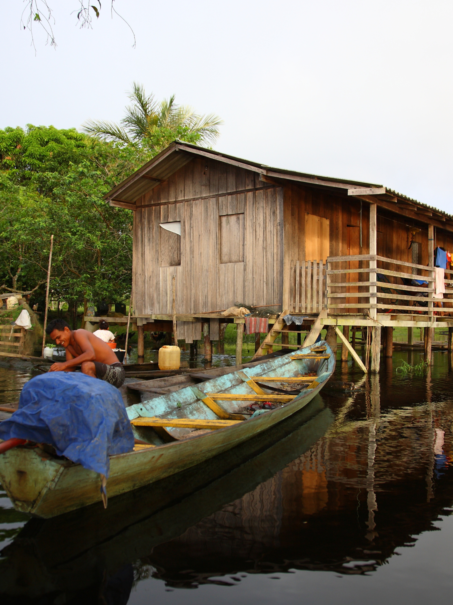 A person paddles a boat. In the foreground, there is a wooden shack on stilts.