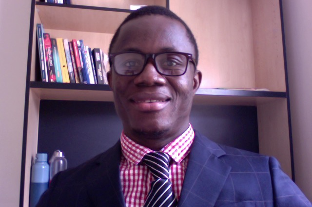 A man with dark skin, short hair and glasses smiles, with a bookshelf in the background