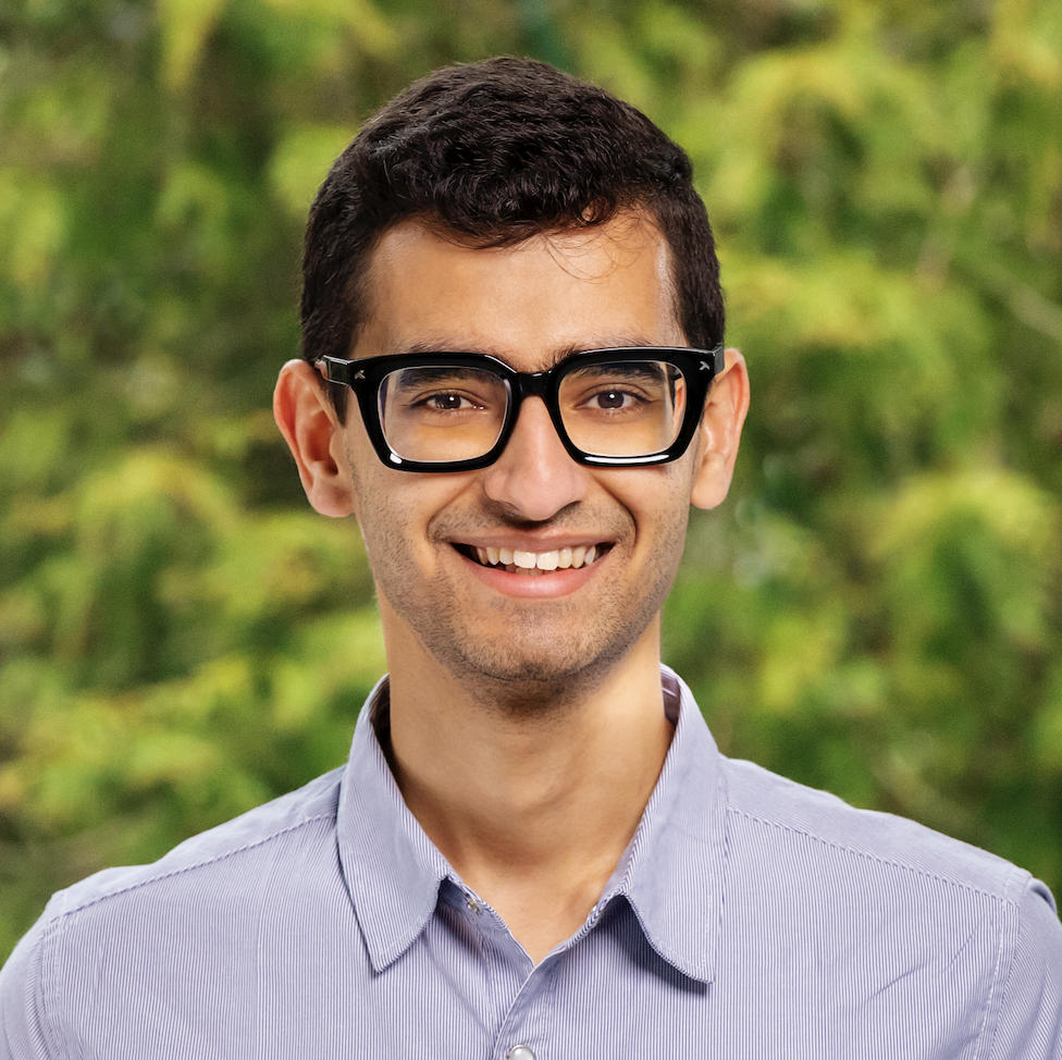 a man with short hair and glasses stands against a green natural background