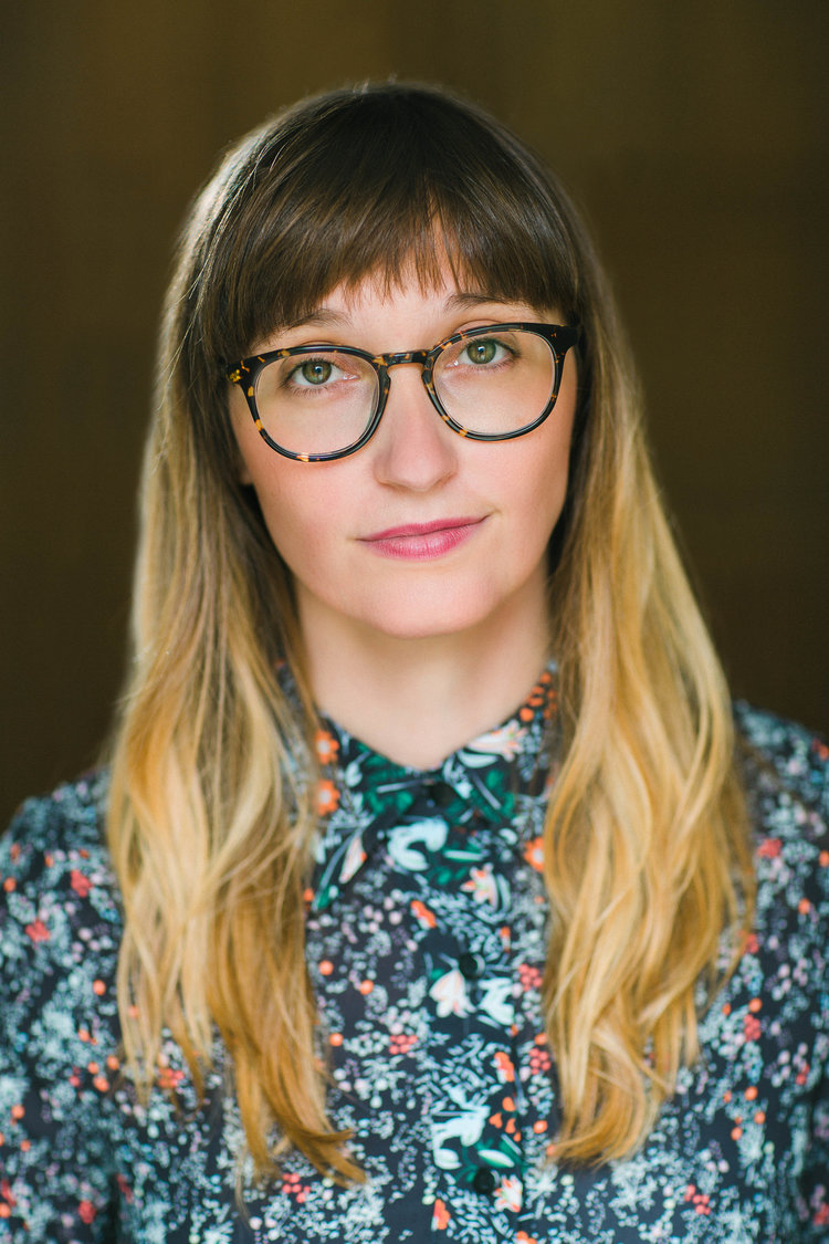 A woman with long hair, bangs, and glasses wears a blue patterned shirt
