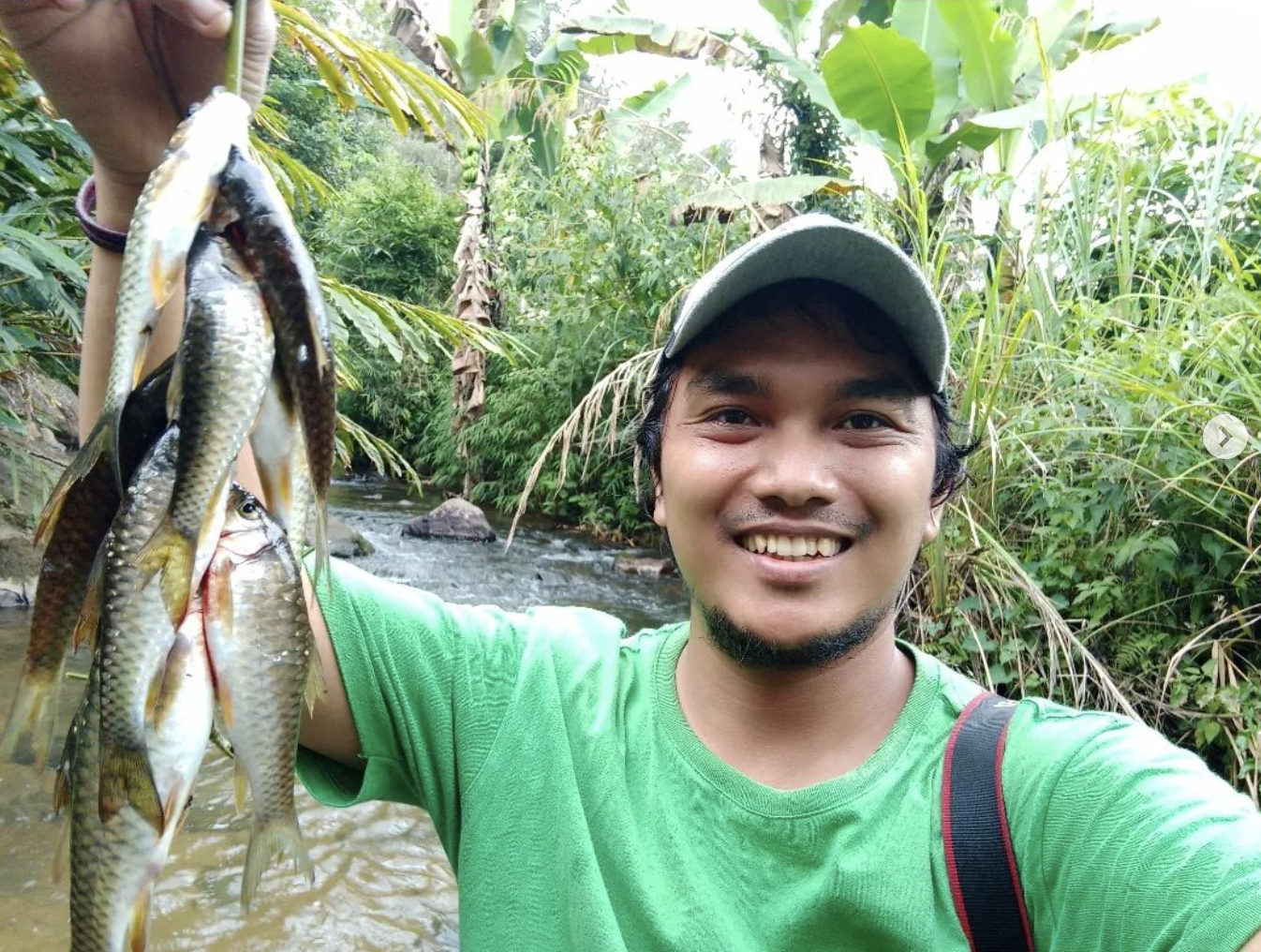 A man wearing a green shirt flashes his teeth as he holds up a bunch of fish in the air.