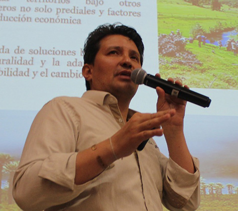 A man wearing a light button-down shirt holds and speaks into a microphone with slides projected behind him