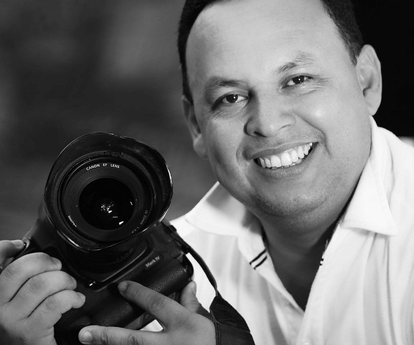 A man gleefully smiles at the camera while holding a camera in his hands.