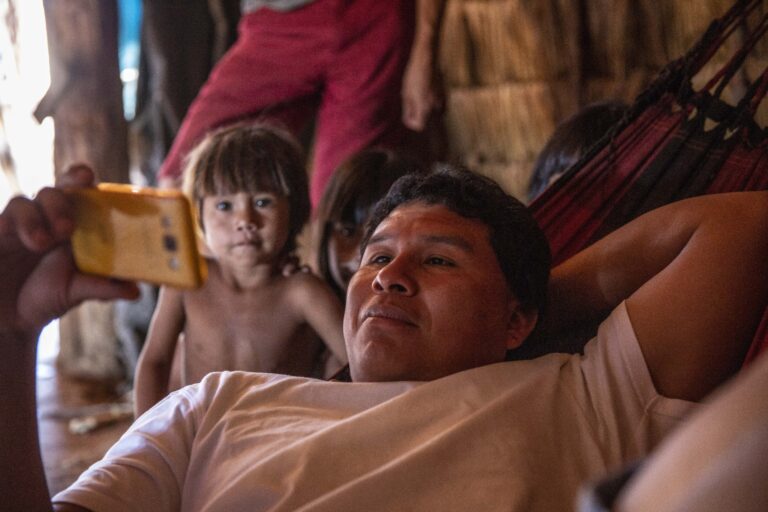 an Indigenous Kalapalo man lies in a hammock holding up a smartphone with a young child next to him