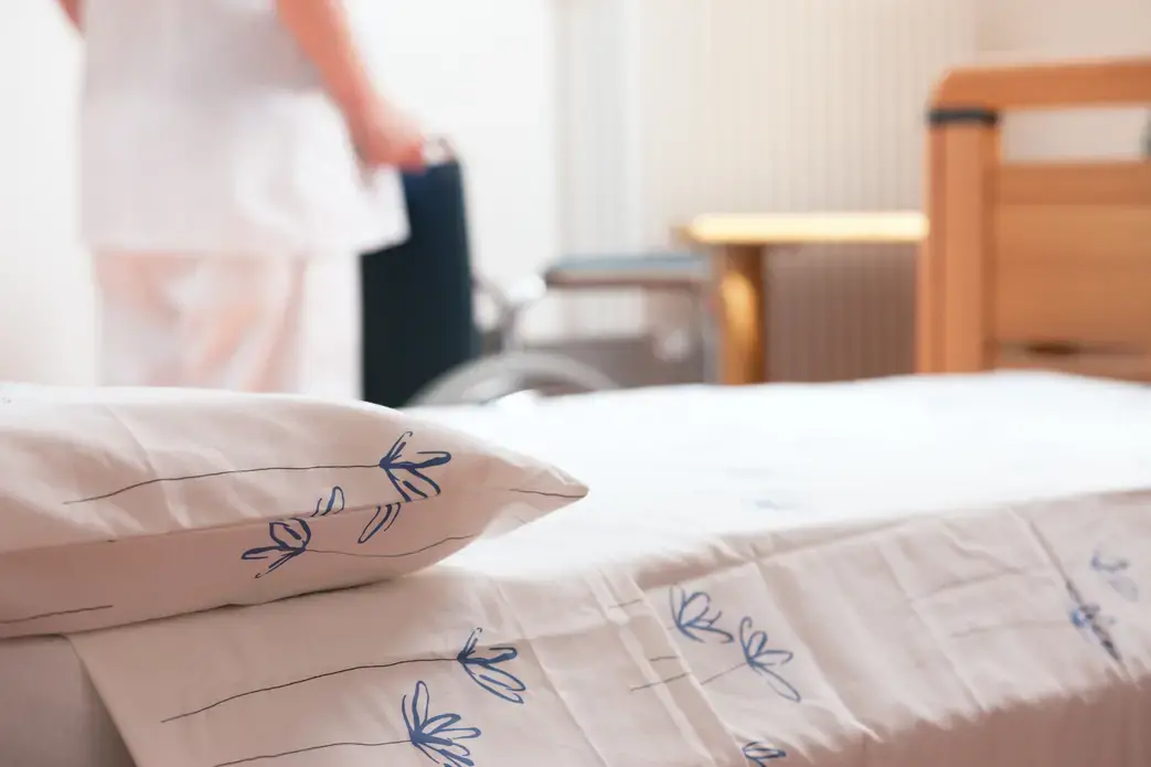 Bed at nursing home with nurse and wheelchair in background. Image by Stokkete / Shutterstock.com. Undated.