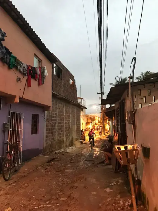 Image of a narrow street at dusk in a community on the outskirts of Recife, Pernambuco. There is an unpaved dirt road with small homes packed close together on either side. A teenager sits on a stoop in the shadows, while a young boy can be seen riding his bike down the street. Image by Poonam Daryani. Brazil, 2017.