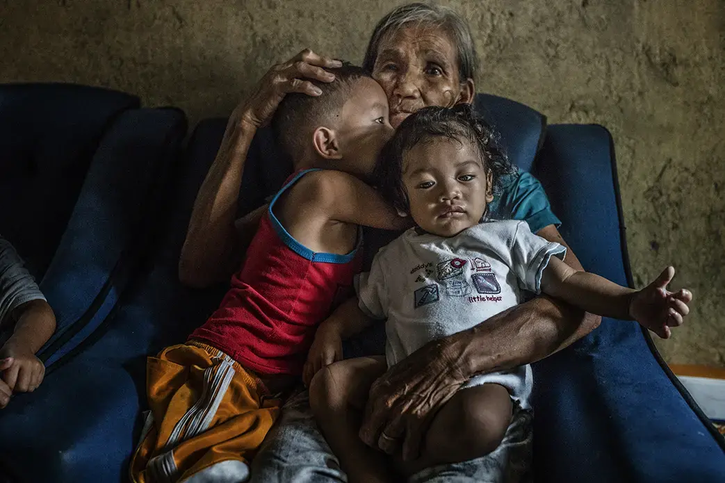 Remy Fernandez, 84-years-old, holds her grandchildren that she is raising, there are seven in all, because her son, Constantino de Juan, a methamphetamine user, was killed by masked men and the mother is in prison due to a drug arrest. The chair in which she sits has a hole in it after it passed through his body. Baby RJ was born in prison. Constantino, upon seeing the masked assassins, instructed CJ, 5-years-old and wearing the red tank top, to take care of his siblings because he knew he was about to be…
