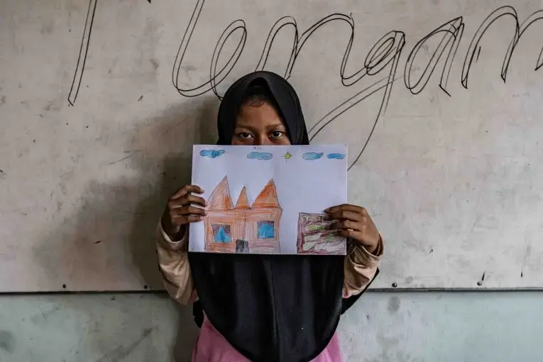 A 10-year old girl holds up her drawing of a home in a government run shelter. The girl is one of the surviving members of the families that carried out a string of ISIS-inspired suicide attacks in the city of Surabaya in May 2018. Image by Jurnasyanto Sukarno. Indonesia, 2018.