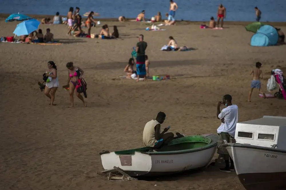 Two migrants sit on a fishing boat as people enjoy the beach in Gran Canaria island, Spain, on Friday, Aug. 21, 2020.&nbsp;Image by Emilio Morenatti/AP Photo. Spain, 2020.