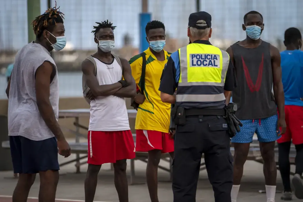 A police officer tells migrants and asylum-seekers to stop playing soccer due to coronavirus restrictions on contact sports in Gran Canaria island, Spain, Tuesday, Aug. 18, 2020. Image by Emilio Morenatti/AP Photo. Spain, 2020.