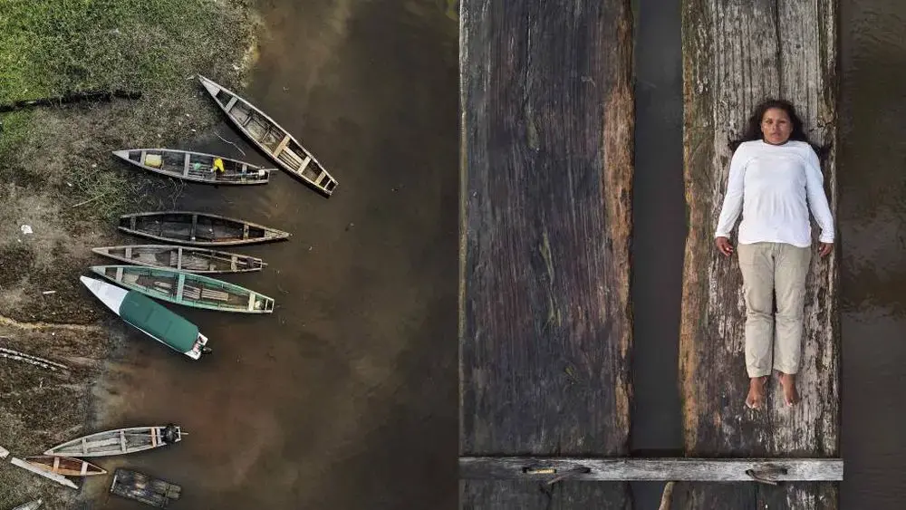 lilia rainforest defenders.jpg To the left, a series of empty wooden canoes stand by the margins of a river. On the right, Lilia Isolina Java Tapayuri lies down on a plank of wood, looking directly at the camera, captured from an aerial shot. She is wearing a white long sleeved shirt, khaki-colored pants, and no shoes.