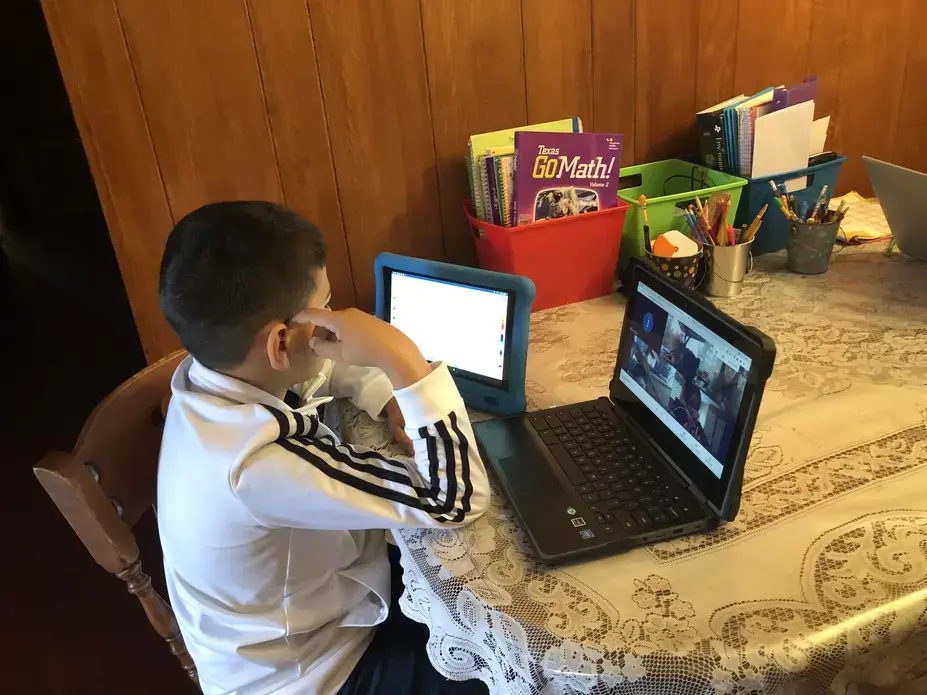 A young boy sits at a kitchen table with a tablet and laptop in front of him. On the laptop, he has a Zoom meeting open with four other attendees. On the table, there are three cups holding writing utensils and red, green, and blue containers holding notebooks and papers.