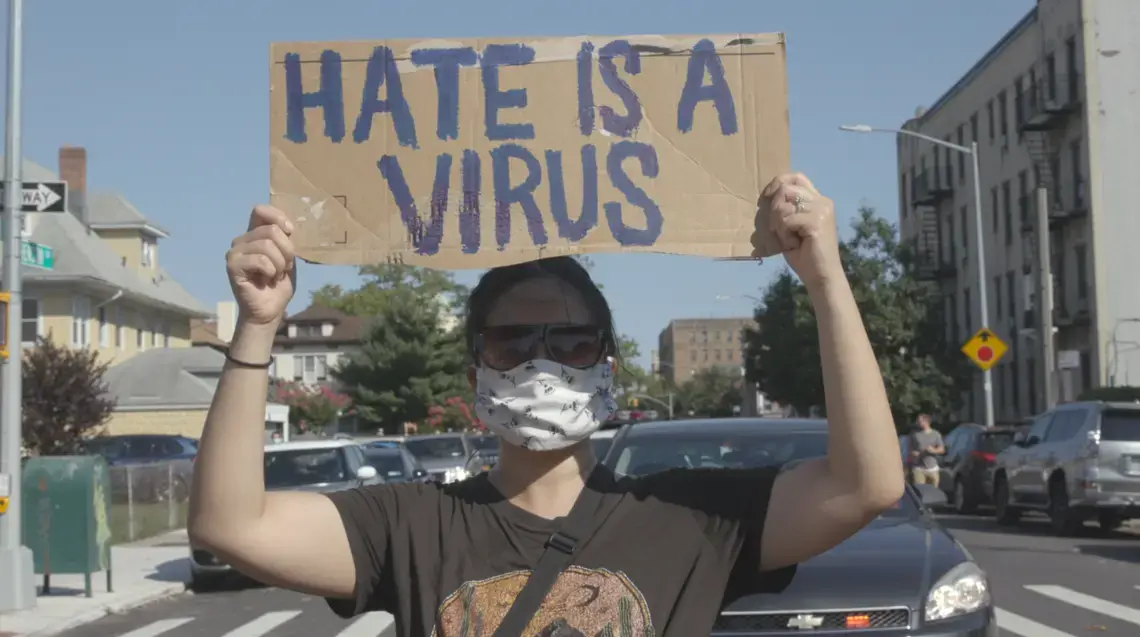 A masked person holds a sign that reads 'hate is a virus'
