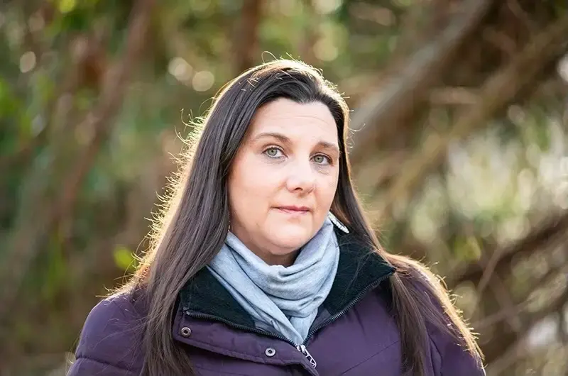 A woman wears a scarf and purple coat in front of trees