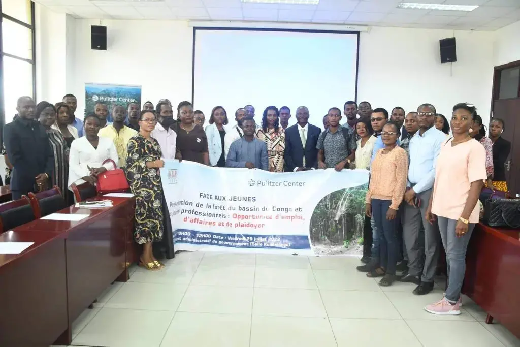 group photo in a conference room. There is a projector and the group together holds a sign that in French reads "Talks with professionals on the Congo Basin" with a Pulitzer Center logo.