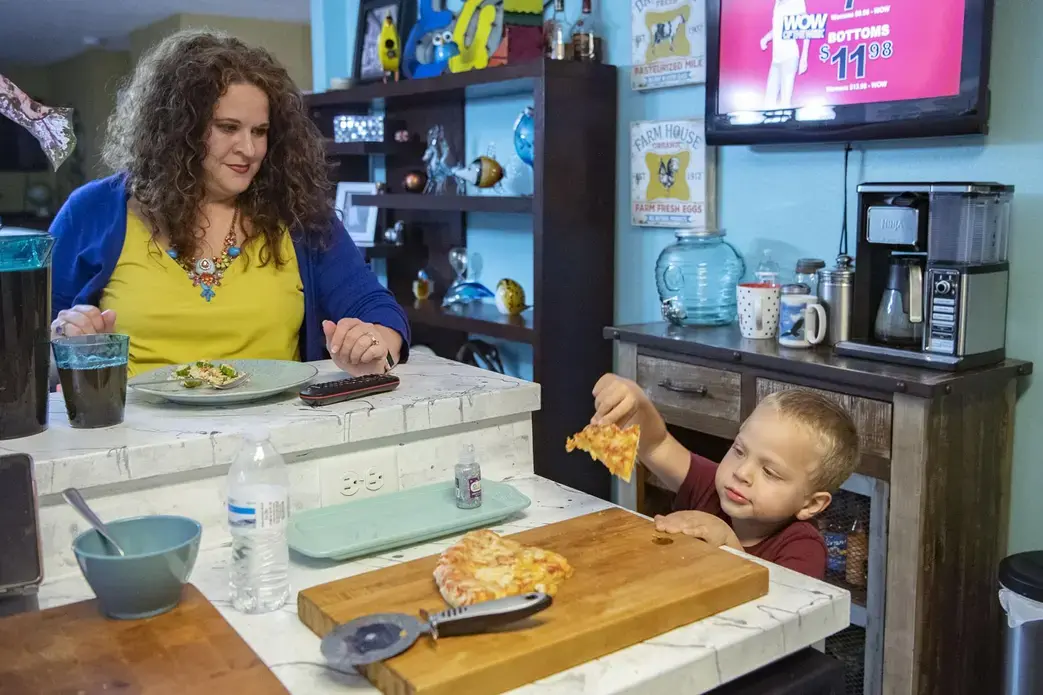 Crystal Camacho finishes her dinner at the kitchen counter as her son, Elias, 4, grabs a second helping of pizza on June 30, 2020 at their home in Swannanoa. Before finding a new day care for Elias after the one he was attending closed, Camacho and her husband had to take turns staying home half days or take the kids to work with them. Image by Angeli Wright/Asheville Citizen Times/North Carolina News Collaborative. United States, 2020.</p>
<p>