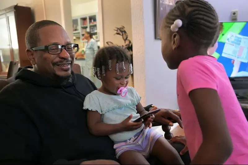 Cecil Burton rocks his granddaughters Chasity Shufford, 1, and Katelyn Ellis, 6, during a family gathering while their mother, Keisha, cooks fried chicken in the kitchen. Burton, 54, said child support once garnished so much of his paycheck he didn't have enough to live on. His coworkers took up a collection for him. Image by Karl Merton Ferron / Baltimore Sun. United States, 2019.<br />
