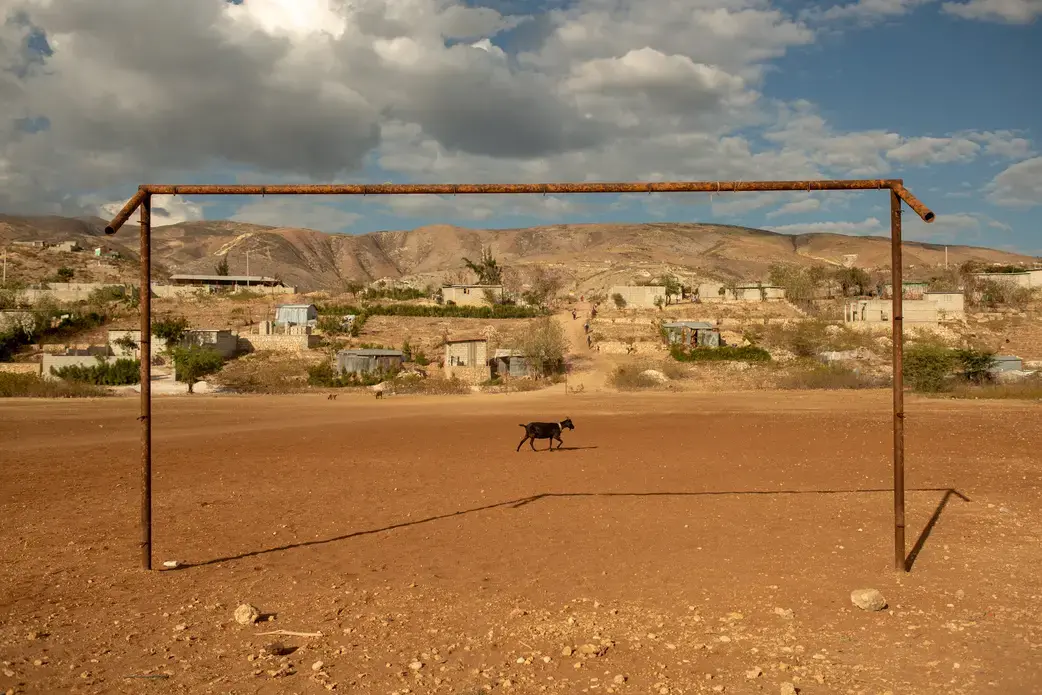 A goat wanders through the community soccer field in the Canaan 1 neighborhood. Local leaders have gone to great lengths to protect the choice piece of land that the pitch is on – a rare flat spot in a hilly region – hoping one day to build a soccer stadium here. Community leaders have asked newcomers who have tried to build on the site to find plots further up the hill. Image by Allison Shelley. Haiti, 2019.