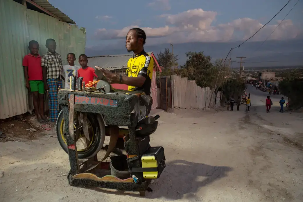 Gemps Rosembert, 31, sharpens knives with a portable machine he has made himself that relies on a pedal driven motorcycle tire to spin the grinding wheel in the Canaan 4 neighborhood. 'I charm my customers with music,' says Rosembert, who is partial to reggae and has named the machine 'File Mix.' He sharpens to a curated musical selection he plays from a boombox. Image by Allison Shelley. Haiti, 2019.