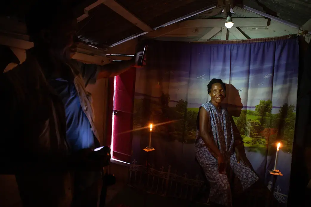 Photographer Charmant Goutier, 46, photographs Marie Micheline Josia Naissance, 54, in his photo studio in the Canaan 4 neighborhood. Goutier moved his family to the neighborhood on June 25, 2011. When the power stays off for awhile and he can't recharge his lights, he relies on candles and a cell phone flashlight to help him focus on his subjects until his on-camera flash can fire. Image by Allison Shelley. Haiti, 2019.