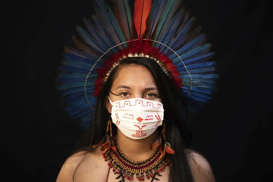 Samela, 23, of the Sateré Mawé indigenous ethnic group, poses for a portrait wearing the traditional dress of her tribe and a face mask amid the spread of the new coronavirus in Manaus, Brazil, Wednesday, May 27, 2020. Samela, whose mother Sonia Vilacio recovered from symptoms consistent with COVID-19 using natural remedies at home, is the first member of her family to attend university, where she studies biology. Image by Felipe Dana / AP Photo. Brazil, 2020.