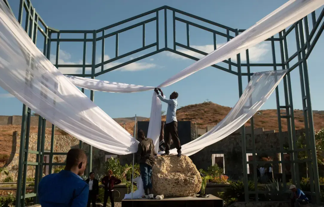 A man ties decorative fabric together at the St. Christophe memorial. In preparation for the annual wreath laying ceremony on the ninth anniversary of the earthquake. Image by Allison Shelley. Haiti, 2019.