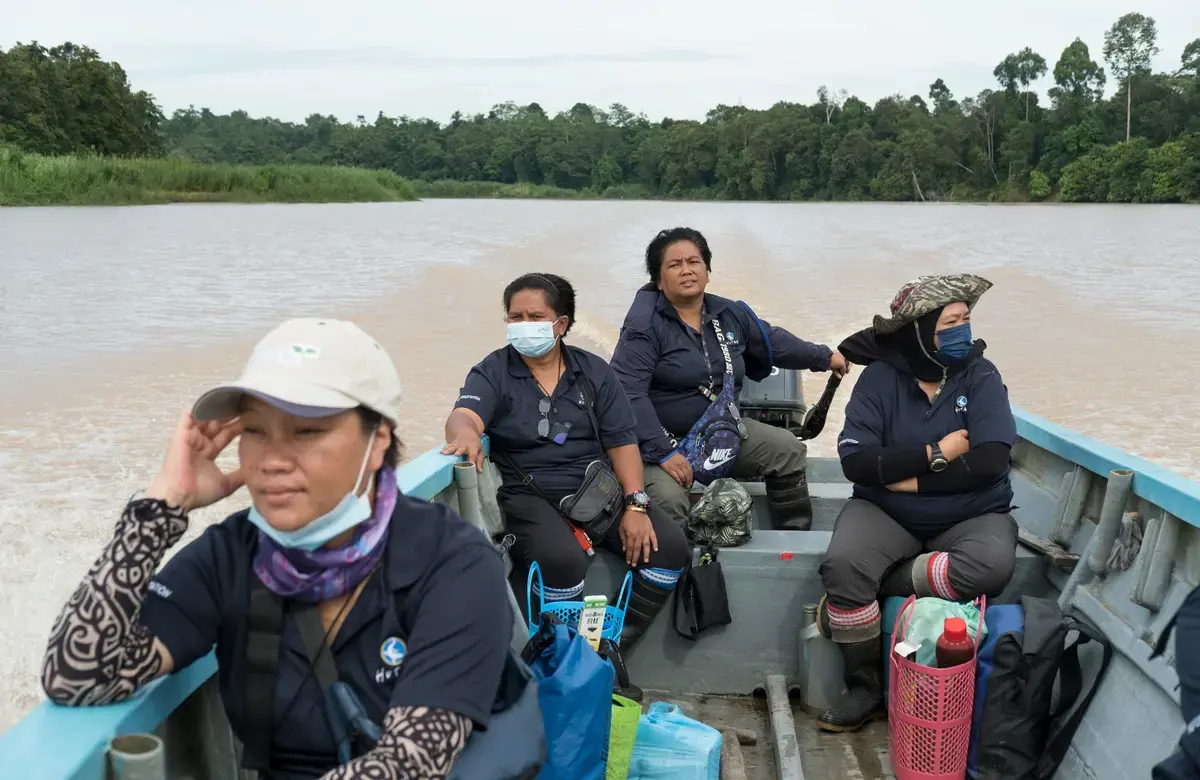 A group of women sit in a boat as it rides down a gray river lined with trees
