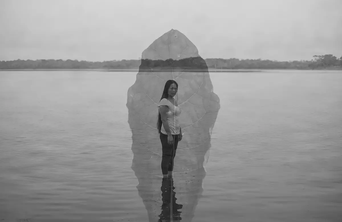 A double exposed photograph of a woman standing in a body of water looking at the camera with a single leaf superimposed over her body