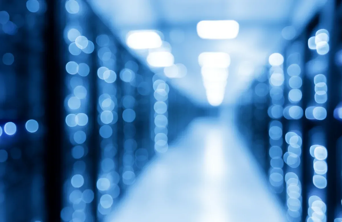 A blurred image of a dimly lit datacenter hallway showing out of focus vertical stacks of lights emitted by computer processors.