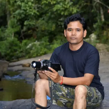 Portrait of Leng Ouch holding a camera and sitting on a rock next to a lake with trees behind him.
