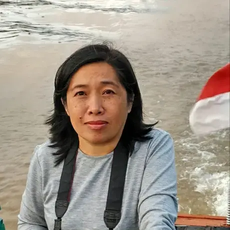 Febrianti.jpg A person wears a grey long sleeve shirt and is carrying a camera. In the foreground there is a beach with ocean waves.