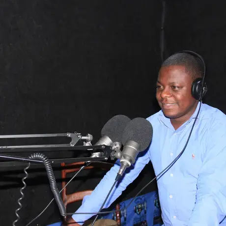 A person wears a blue shirt and stands over a microphone. He is also wearing headphones and is in a radio station.
