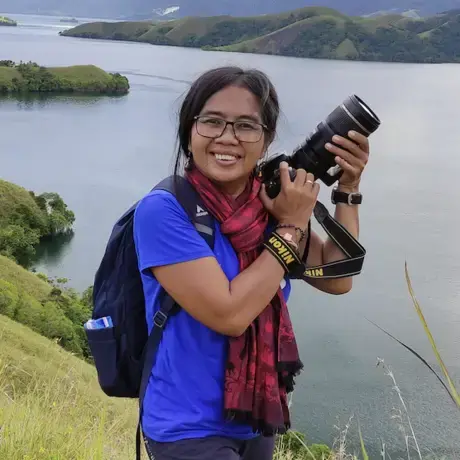A woman holds a camera in her hands. She wears glasses, a purple shirt and jeans.