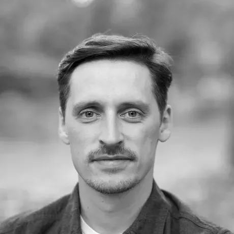 A black and white photo of a man with short dark hair wearing a white tshirt and a dark button down