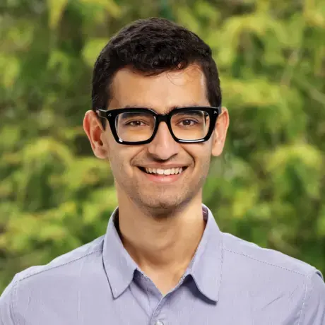 a man with short hair and glasses stands against a green natural background
