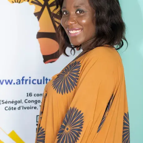 A woman smiles clad in a mustard dress against a flex standee with a map of Africa on it.
