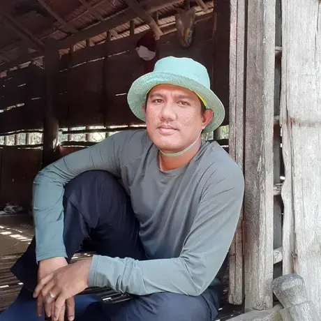 Suryadi sits on a threshold wearing a bucket hat.