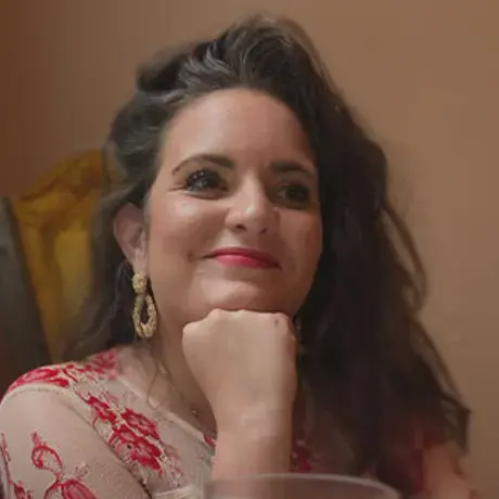 A woman with long dark wavy hair rests her chin on her fist while seated at a table