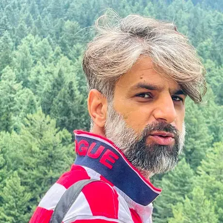 A man with grey hair and a beard wears a red and white striped shirt against a wooded background