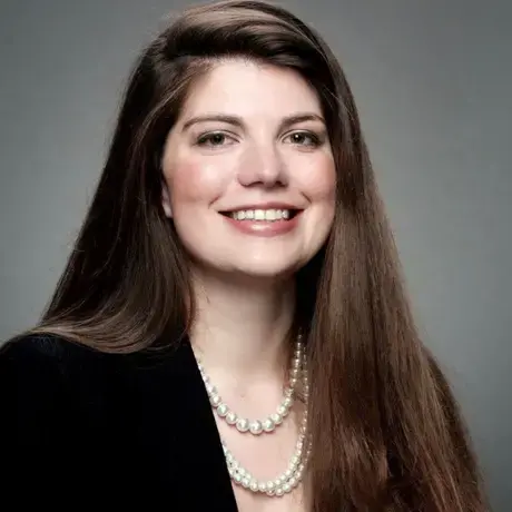 A woman with long dark hair wearing a black blazer against a grey background