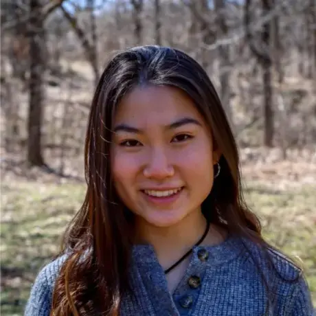 Headshot of a woman with long dark hair wearing a grey sweater against a wooded background