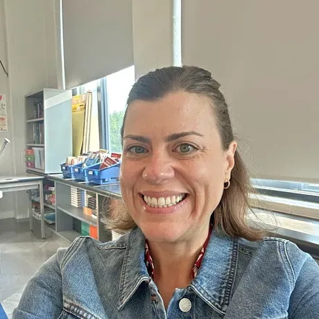 Jodi Hoard is a white woman with light brown hair pulled away from her face. She is standing in a classroom and smiling into the camera.