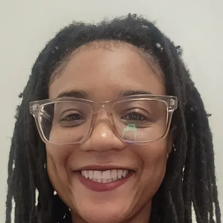 Nkomo is a Black woman with dreadlocks and glasses smiling into the camera.