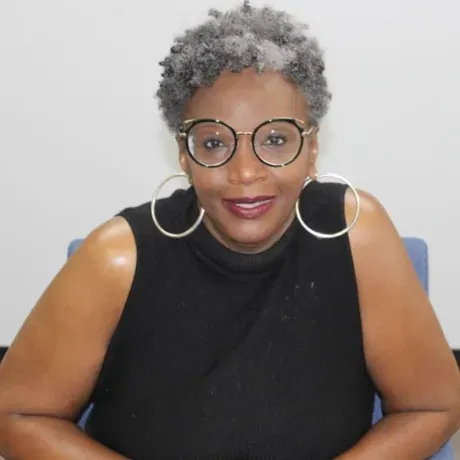 A photo of Valencia Abbot. A Black woman with glasses, hoop earrings, and a short gray afro smiles into the camera.