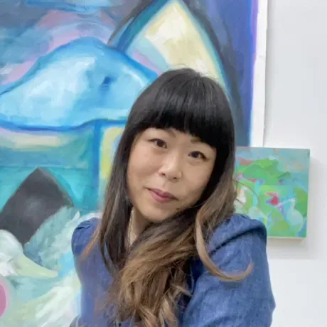 Jane Lawrence is a Asian woman with long brown black hair. She stands in front of artwork smiling into the camera.