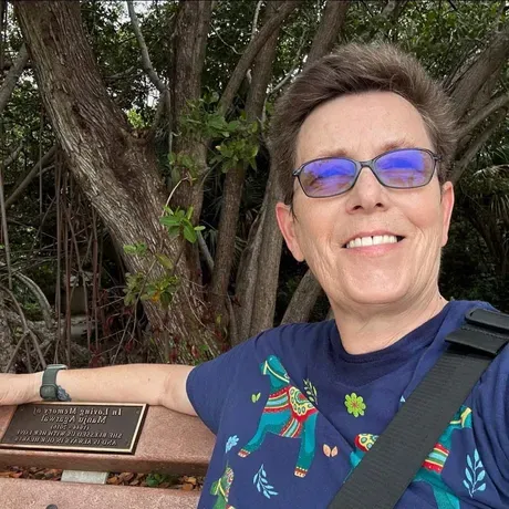 A selfie of Kris Erickson. She is wearing a blue shirt, sunglasses, and smiling into the camera.